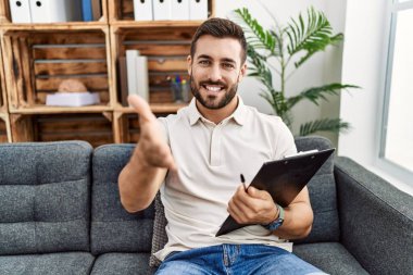 Handsome hispanic man holding clipboard working at psychology clinic smiling friendly offering handshake as greeting and welcoming. successful business. 