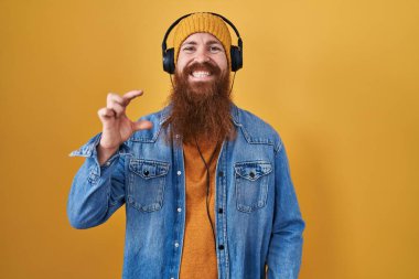 Caucasian man with long beard listening to music using headphones smiling and confident gesturing with hand doing small size sign with fingers looking and the camera. measure concept. 