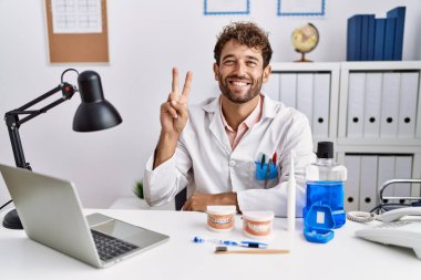 Young hispanic dentist man working at medical clinic showing and pointing up with fingers number two while smiling confident and happy. 