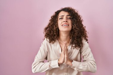 Hispanic woman with curly hair standing over pink background begging and praying with hands together with hope expression on face very emotional and worried. begging. 