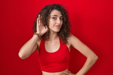 Hispanic woman with curly hair standing over red background smiling with hand over ear listening an hearing to rumor or gossip. deafness concept. 