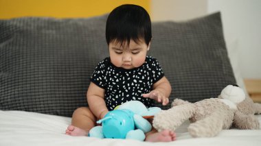 Adorable hispanic baby playing with dolls sitting on bed at bedroom