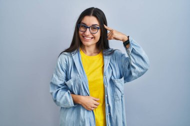 Mavi arka planda duran genç İspanyol kadın gülümseyerek tek parmağıyla kafasını işaret ediyor, harika bir fikir ya da düşünce, iyi bir hafıza. 