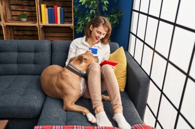 Genç beyaz bir kadın dokunmatik ped ve kredi kartı kullanıyor. Evde köpekle birlikte kanepede oturuyor.