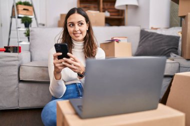 Young beautiful hispanic woman using laptop and smartphone sitting on floor at new home