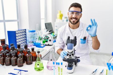 Handsome hispanic man working at scientist laboratory doing ok sign with fingers, smiling friendly gesturing excellent symbol 