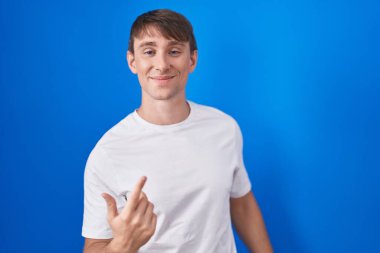 Caucasian blond man standing over blue background beckoning come here gesture with hand inviting welcoming happy and smiling 