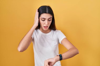 Young beautiful woman standing over yellow background looking at the watch time worried, afraid of getting late 