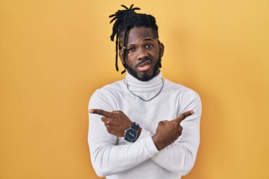 Sarı arka plan üzerine balıkçı yaka kazak giyen Afrikalı adam iki tarafı da parmaklarıyla işaret ediyor. Farklı yönler farklı. 