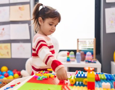 Adorable hispanic girl playing with construction blocks standing at kindergarten