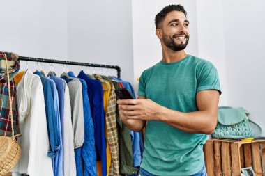 Genç Arap müşteri akıllı telefon kullanıyor giyim mağazasında elbise rafının yanında duruyor.