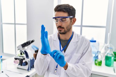 Young hispanic man wearing scientist uniform and gloves at laboratory