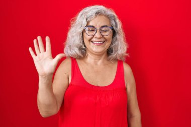 Middle age woman with grey hair standing over red background showing and pointing up with fingers number five while smiling confident and happy. 
