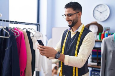 Genç Arap terzi, giyim fabrikasında akıllı telefon kullanarak gülümsüyor.