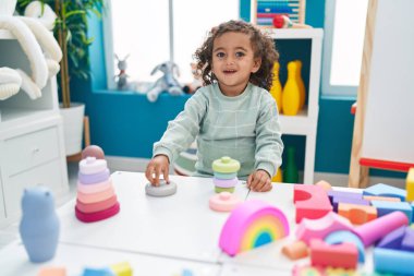 Adorable hispanic girl playing with construction blocks standing at kindergarten