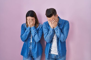 Young hispanic couple standing over pink background with sad expression covering face with hands while crying. depression concept. 