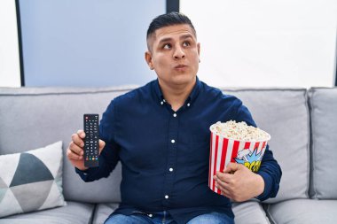 Hispanic young man eating popcorn using tv control smiling looking to the side and staring away thinking. 