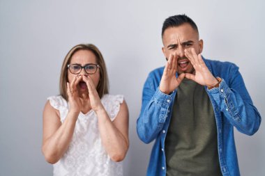 Hispanic mother and son standing together shouting angry out loud with hands over mouth 