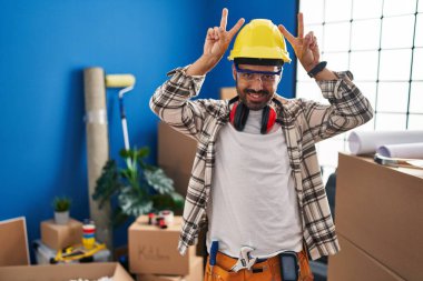 Sakallı, evde çalışan, komik ve deli gibi poz veren, parmakları tavşan kulaklı, neşeli gülümseyen genç İspanyol bir adam. 