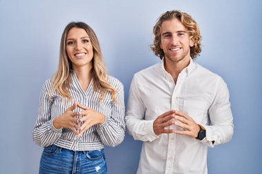 Young couple standing over blue background hands together and fingers crossed smiling relaxed and cheerful. success and optimistic 