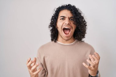 Hispanic man with curly hair standing over white background crazy and mad shouting and yelling with aggressive expression and arms raised. frustration concept. 