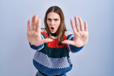 Young hispanic girl standing over blue background doing stop gesture with hands palms, angry and frustration expression 