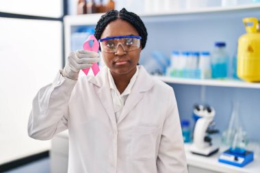 African american woman working at scientist laboratory holding pink ribbon thinking attitude and sober expression looking self confident 