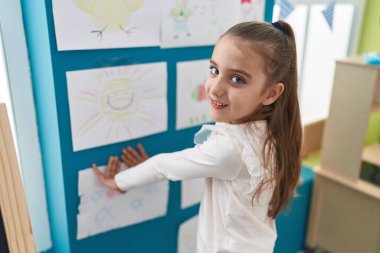 Adorable hispanic girl preschool student hanging draw on wall at kindergarten