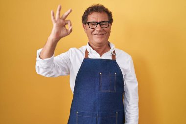 Middle age hispanic man wearing professional cook apron smiling positive doing ok sign with hand and fingers. successful expression. 