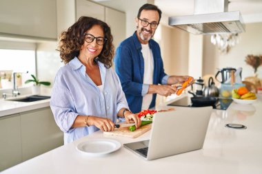 Orta yaşlı İspanyol çift mutfakta yemek pişiriyor ve laptop kullanıyor.
