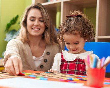 Teacher and toddler playing with maths puzzle game sitting on table at kindergarten