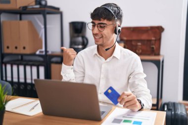 Young hispanic man working using computer laptop holding credit card smiling with happy face looking and pointing to the side with thumb up. 