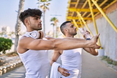 Two hispanic men couple smiling confident stretching and drinking water at street