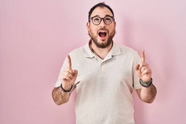 Plus size hispanic man with beard standing over pink background amazed and surprised looking up and pointing with fingers and raised arms. 