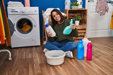 Young beautiful hispanic woman smiling confident holding clean products at laundry room