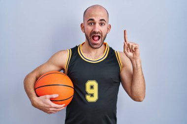 Young bald man with beard wearing basketball uniform holding ball smiling amazed and surprised and pointing up with fingers and raised arms. 