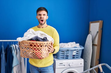 Hispanic man holding laundry basket angry and mad screaming frustrated and furious, shouting with anger. rage and aggressive concept. 