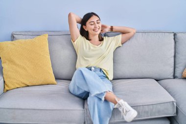 Young hispanic woman relaxed with hands on head sitting on sofa at home