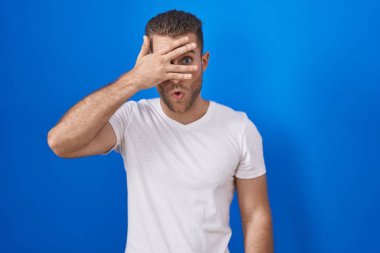 Young caucasian man standing over blue background peeking in shock covering face and eyes with hand, looking through fingers with embarrassed expression. 