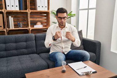 Young hispanic man with beard working at consultation office ready to fight with fist defense gesture, angry and upset face, afraid of problem 