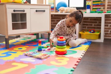 Adorable hispanic girl playing with maths puzzle game sitting on floor at kindergarten