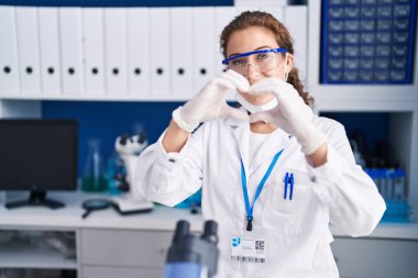 Young caucasian woman working at scientist laboratory smiling in love doing heart symbol shape with hands. romantic concept. 