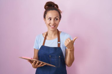 Brunette woman wearing professional waitress apron holding clipboard pointing thumb up to the side smiling happy with open mouth 