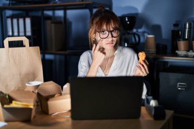 Young beautiful woman working using computer laptop and eating delivery food pointing to the eye watching you gesture, suspicious expression 