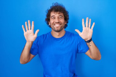 Latin kökenli genç adam mavi arka planda duruyor ve parmaklarını on numarayı gösterip işaret ederken kendinden emin ve mutlu gülümsüyor.. 