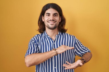 Hispanic man with long hair standing over yellow background gesturing with hands showing big and large size sign, measure symbol. smiling looking at the camera. measuring concept. 