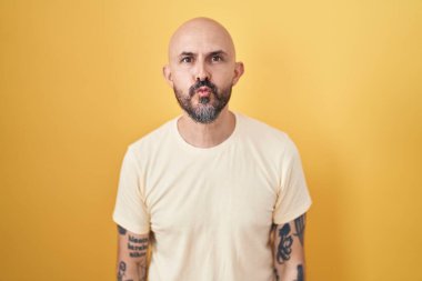 Hispanic man with tattoos standing over yellow background looking at the camera blowing a kiss on air being lovely and sexy. love expression. 