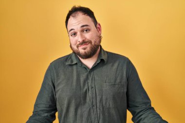 Plus size hispanic man with beard standing over yellow background smiling cheerful with open arms as friendly welcome, positive and confident greetings 