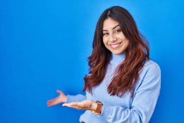 Hispanic young woman standing over blue background inviting to enter smiling natural with open hand 