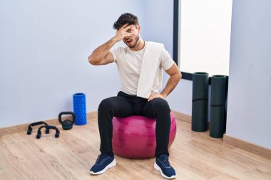 Hispanic man with beard sitting on pilate balls at yoga room peeking in shock covering face and eyes with hand, looking through fingers with embarrassed expression. 